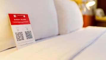 A hotel room with a bed, pillows, a bedside table, and a sign for in-room delivery services.