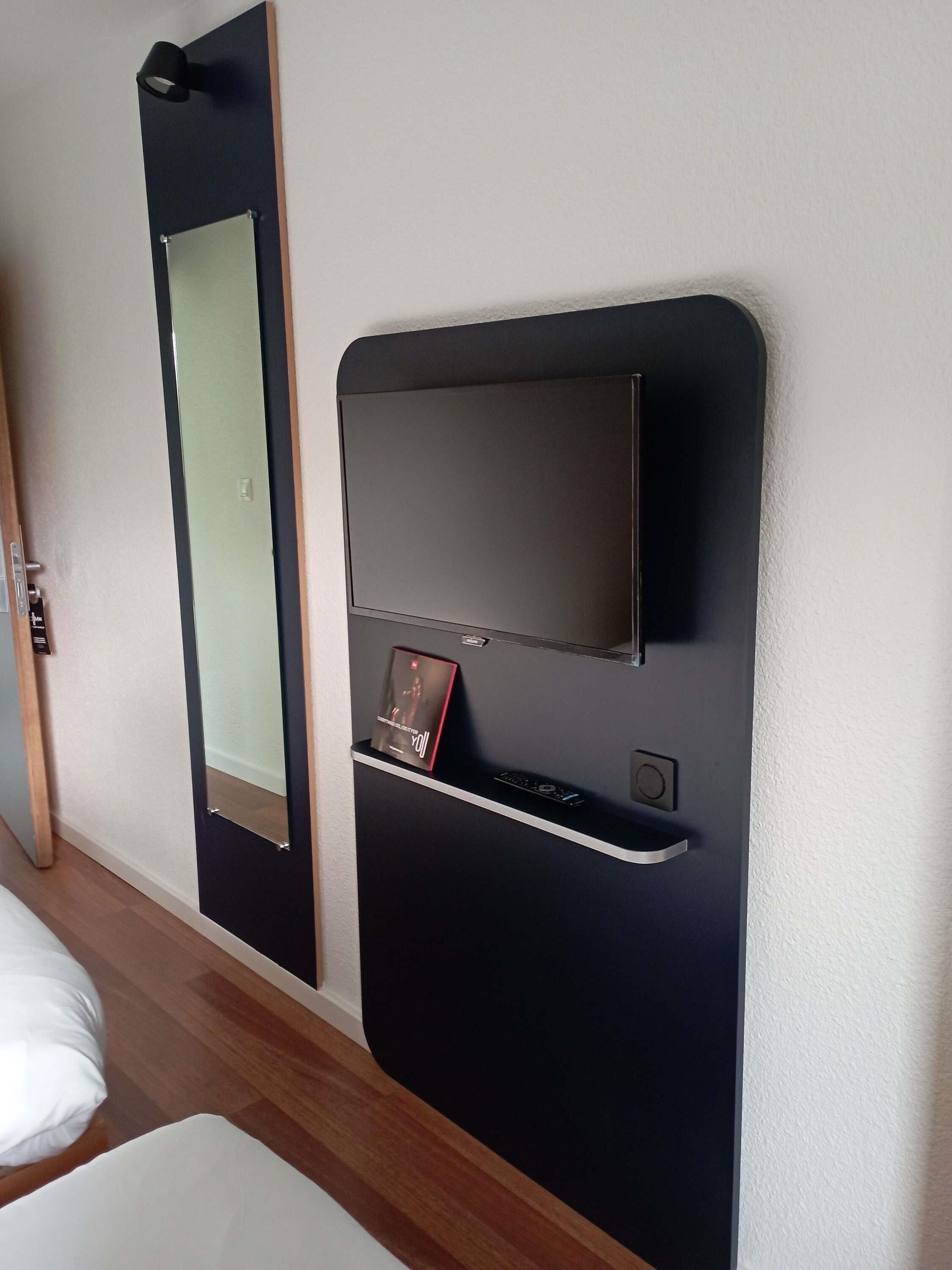 A room with a bed, a mirror, a TV, and a remote control on a stand.