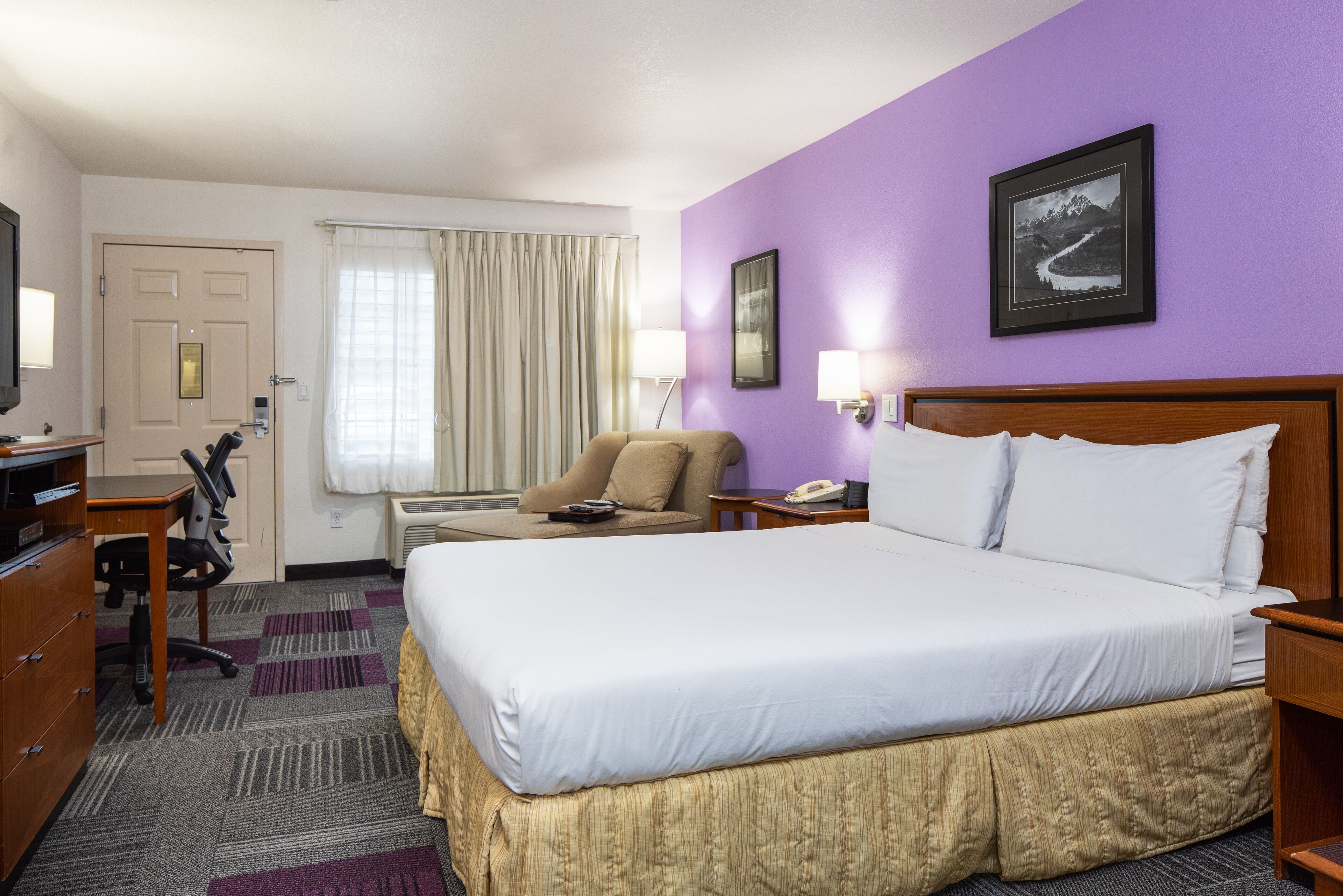 A hotel room with a large bed, a desk with a chair, a television, and a window with curtains.