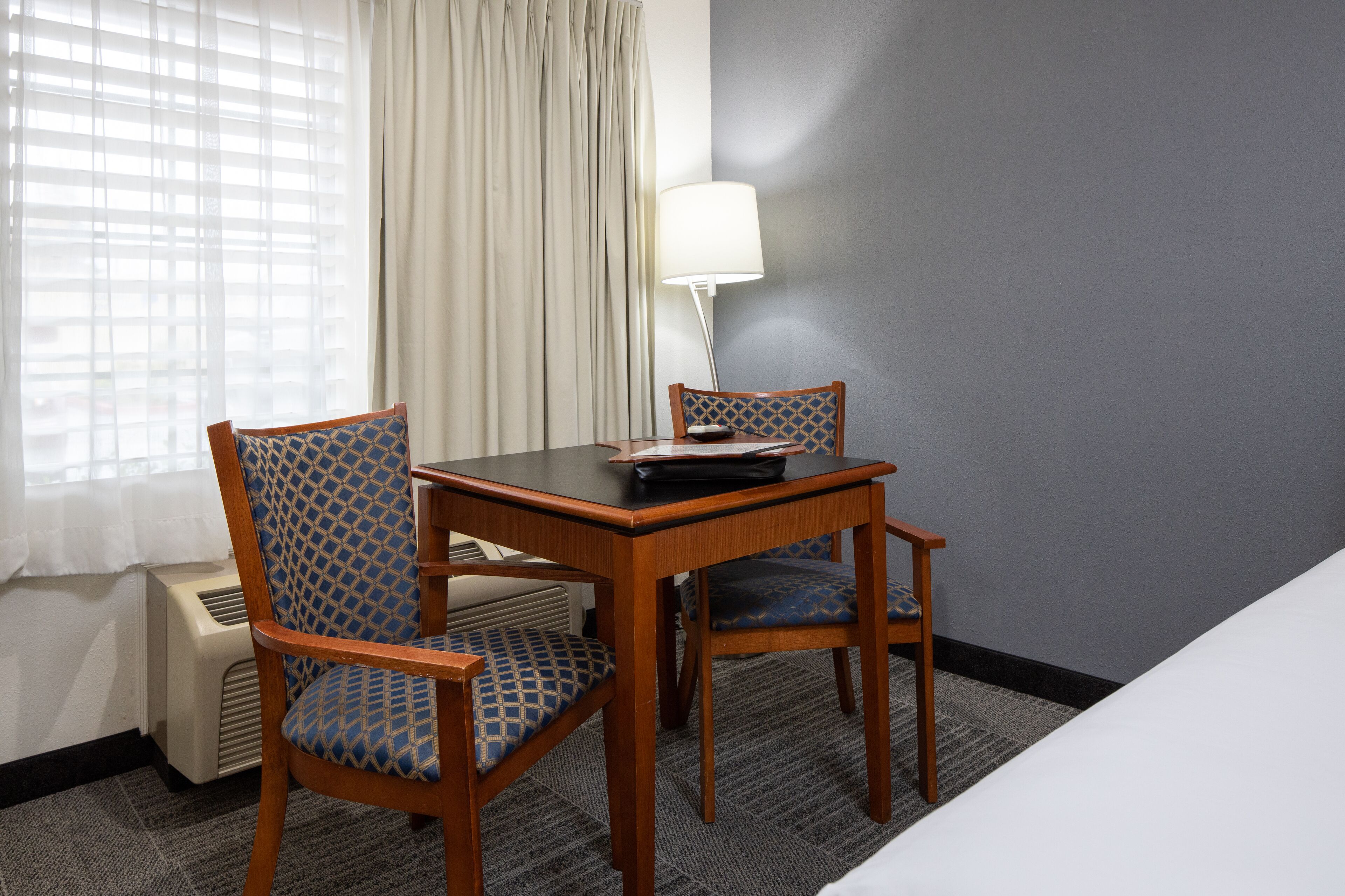 Habitación de hotel con una mesa de madera, dos sillas estampadas, una lámpara y una ventana con cortinas.