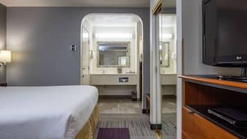 Quarto de hotel com cama, televisão, banheiro com espelho e escrivaninha.