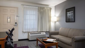 A hotel room with a sofa, a table with a remote control, a treadmill, and a window with curtains. - Atherton Park Inn & Suites (Redwood City)