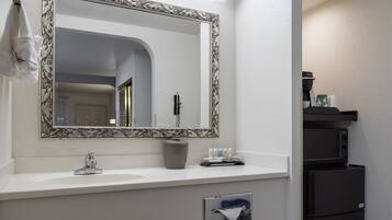 A bathroom with a white sink, a mirror with a decorative frame, a towel hanging on the wall, a small trash bin, and a black refrigerator.