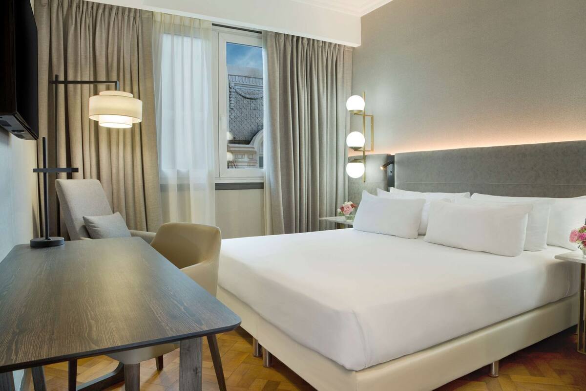 A hotel room with a bed, a desk, a chair, a TV, and a window with curtains.