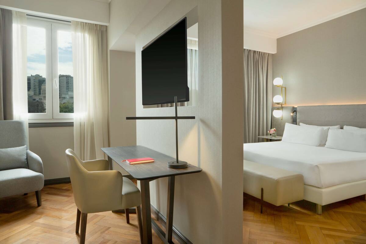 A hotel room with a bed, a desk with a chair, a TV, and a view of buildings outside the window.