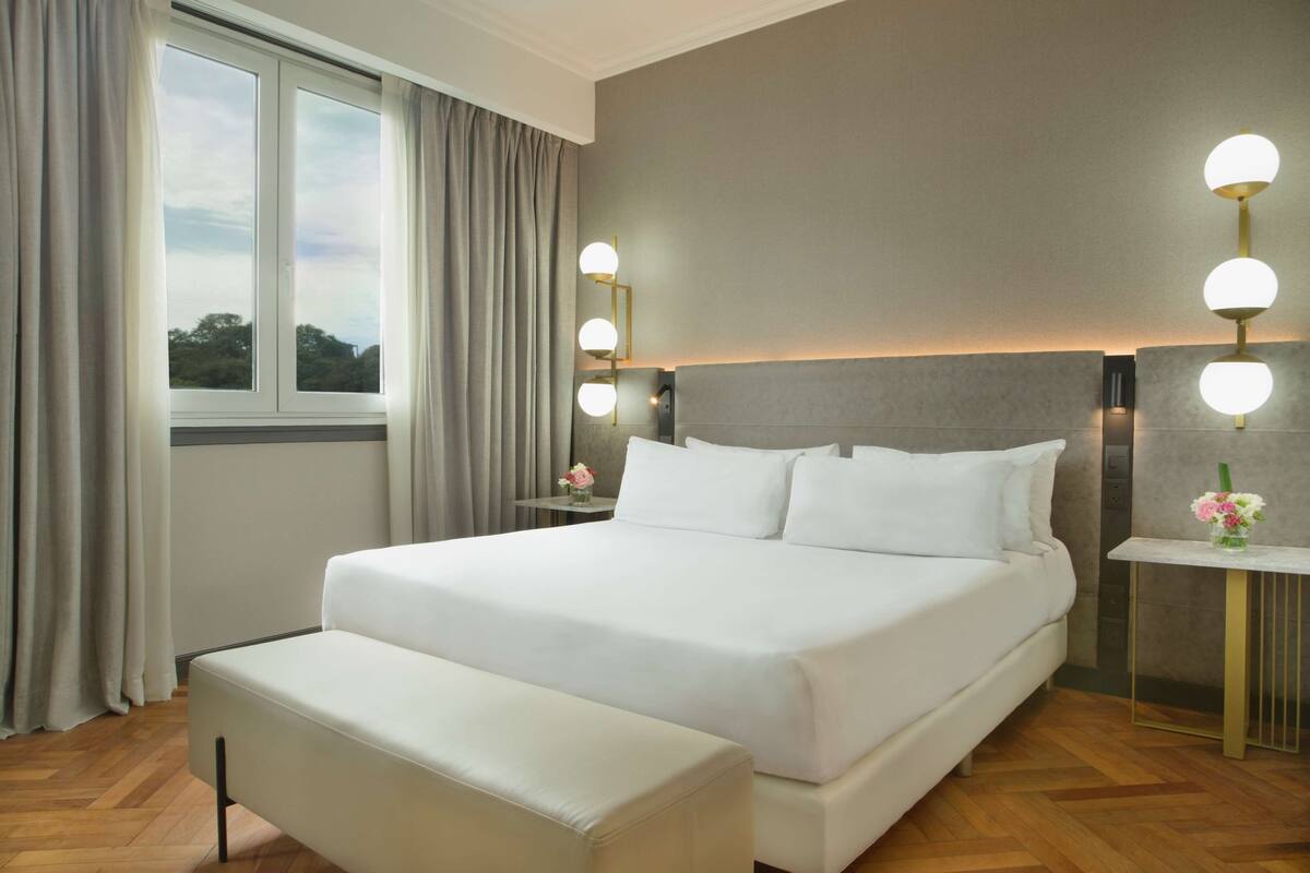 A modern hotel room with a large bed, a bedside table with a lamp, and a view of the outdoors through a window.