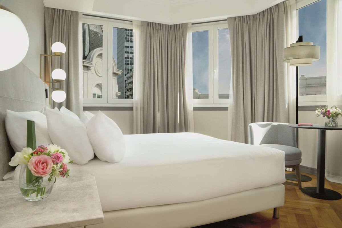 A hotel room with a large bed, a desk with a lamp, a chair, and a view of buildings through the window.