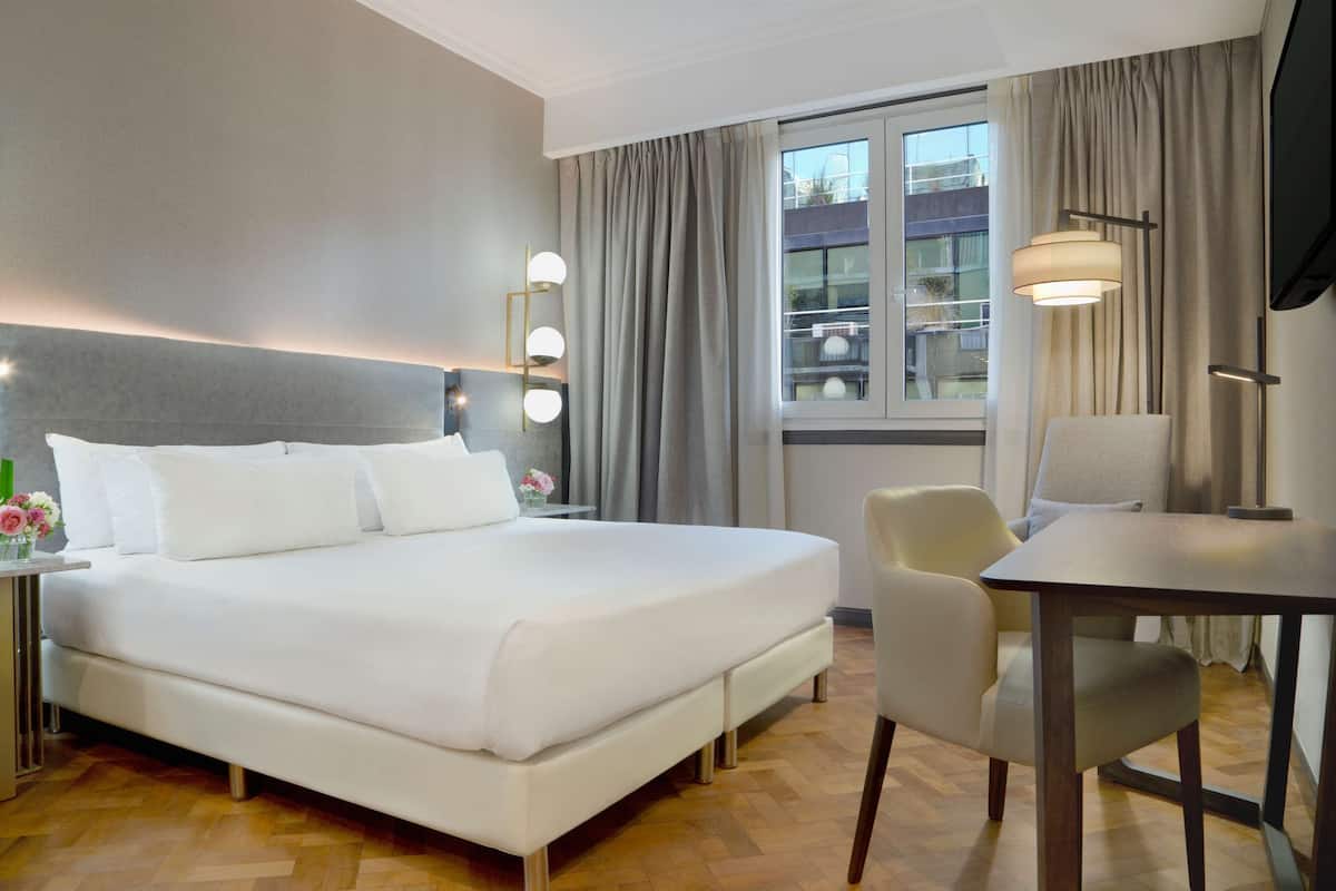 A hotel room with a large bed, bedside table with lights, a chair, a desk, and a window with curtains.