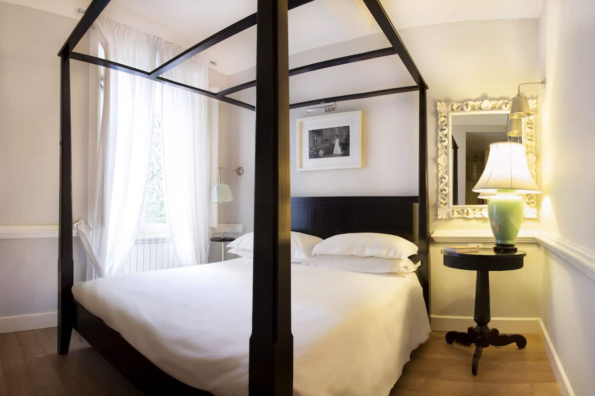 A four-poster bed with white linens, a bedside table with a lamp, and a framed picture on the wall.