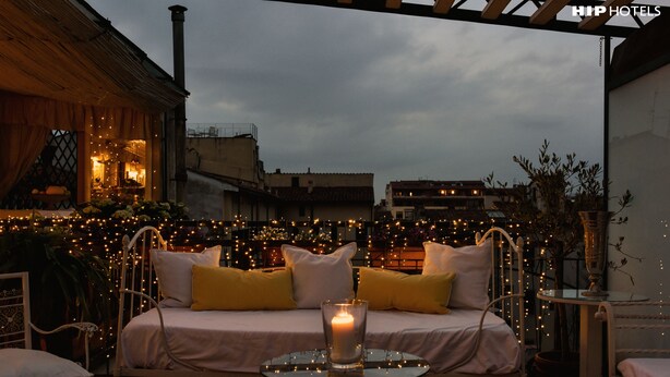 Una zona de estar al aire libre acogedora, con un banco acolchado, una mesa de cristal con una vela y luces colgantes.