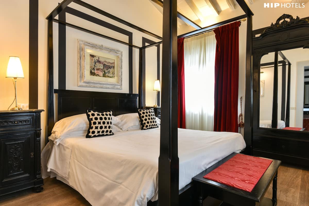 A four-poster bed with white linens and black and white patterned pillows, a bedside table with a lamp, and a red throw on the foot of the bed.