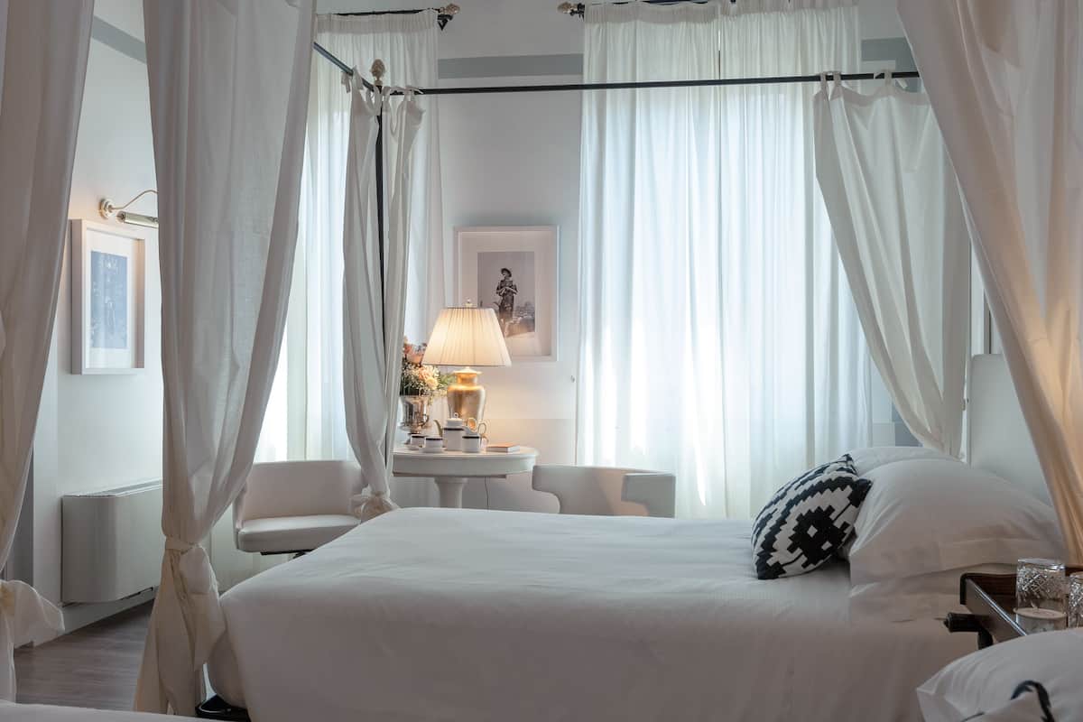 A bedroom with a large bed, white curtains, a bedside table with a lamp, and a chair.