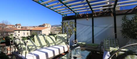 A sunlit outdoor seating area with a glass table, a metal chair, and a sofa with patterned cushions.