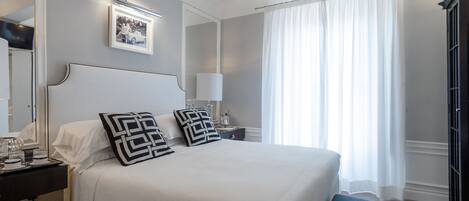 A neatly arranged bedroom with a large bed, a bedside table with a lamp, a mirror, and a framed picture on the wall.