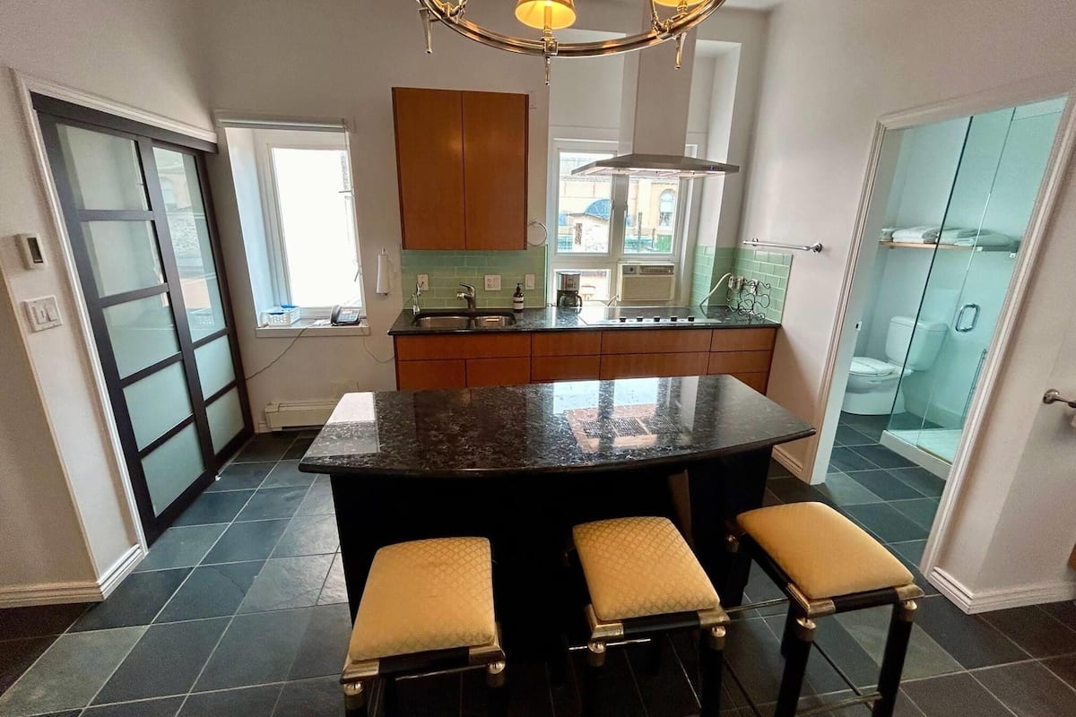 A kitchen with a central island, bar stools, a window, and a glass-enclosed bathroom.