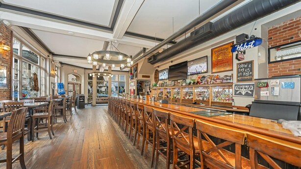 A bar area with wooden bar stools, a long counter, and a brick wall. - Bedford Regency Hotel, Trademark Collection by Wyndham (Victoria)