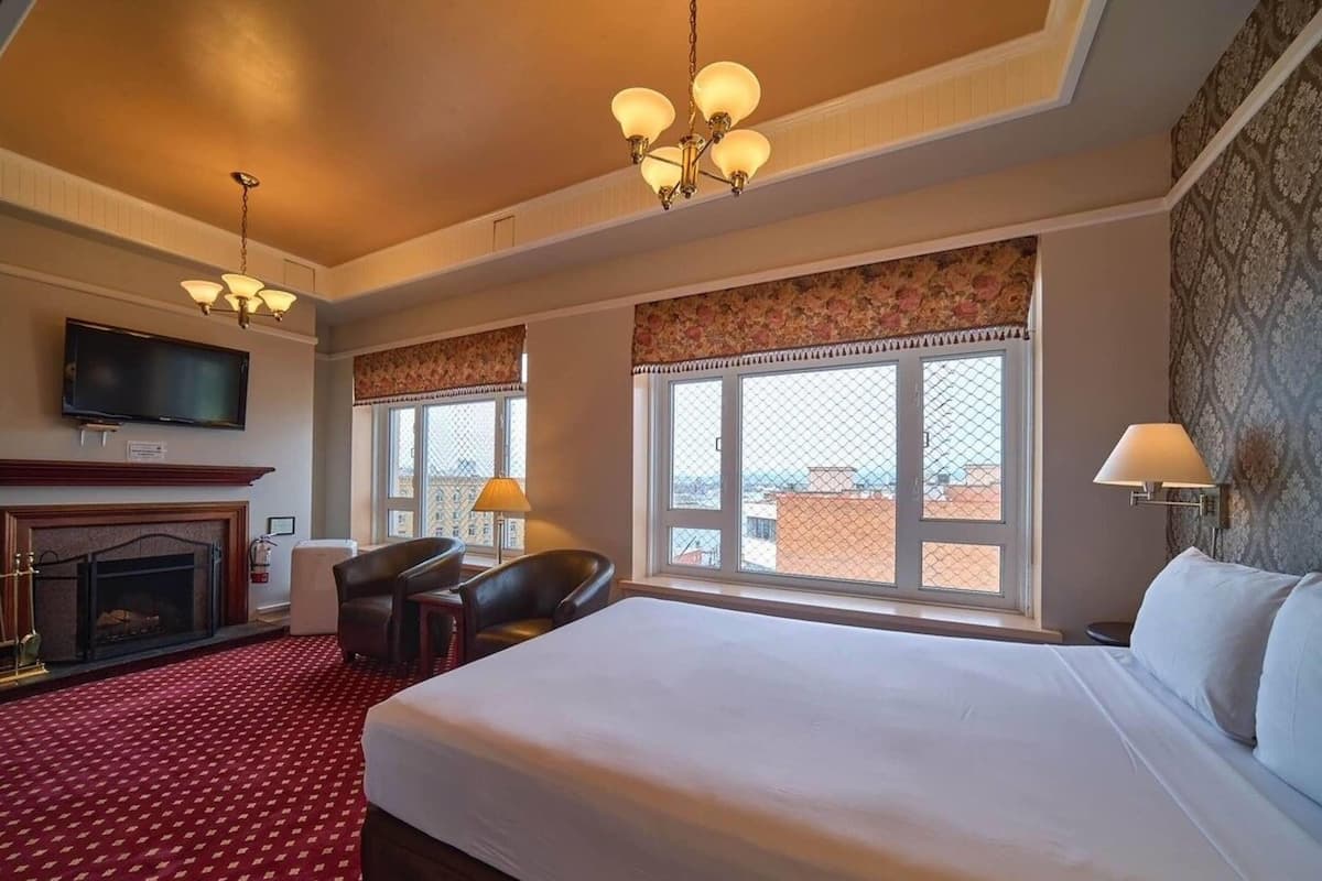 A hotel room with a large bed, a fireplace, a television, and a view of the city through large windows.