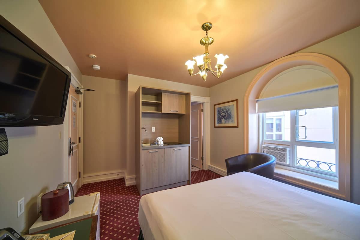 A hotel room with a bed, a TV, a kitchenette, a chair, and a window with a balcony view.