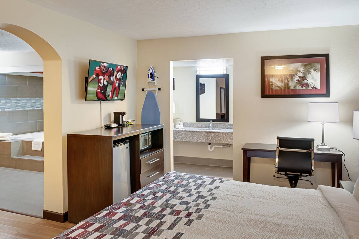 A hotel room with a bed, a desk, a chair, a TV, a microwave, a sink, and a mirror.