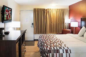 A hotel room with a bed, a desk, a chair, two lamps, a television, and a door. - Red Roof Inn Indianapolis East (Indianapolis)
