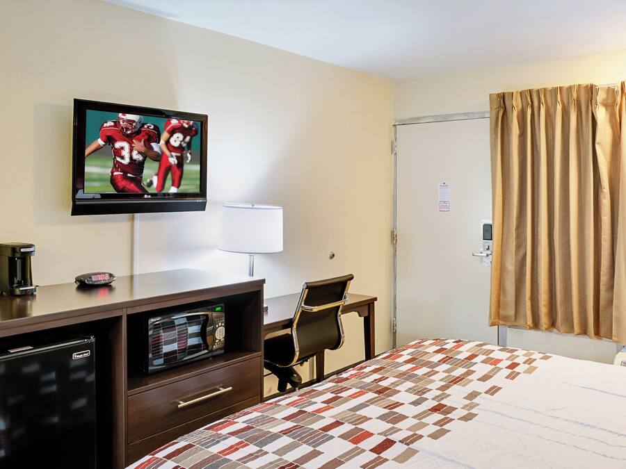 Quarto de hotel com cama, escrivaninha com computador, televisão exibindo uma partida de futebol, cafeteira e relógio.