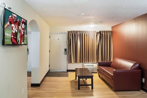 A room with a wooden floor, a brown leather sofa, a small table, a TV mounted on the wall, and a door with a curtain. - Red Roof Inn Indianapolis East (Indianapolis)