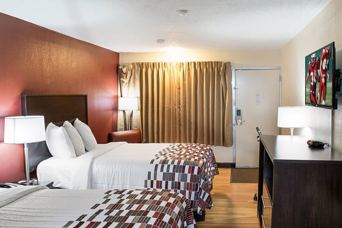 A hotel room with a bed, two bedside tables, a desk, a lamp, a door, and a painting on the wall.