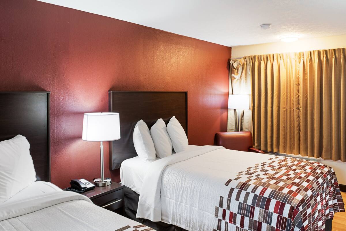 A hotel room with two beds, a nightstand with a lamp, and a window with curtains.