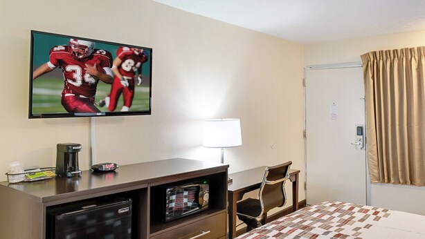 A hotel room with a television mounted on the wall, a desk with a coffee maker, a microwave, a bed with a patterned bedspread, and a door with a curtain. - Red Roof Inn Indianapolis East (Indianapolis)