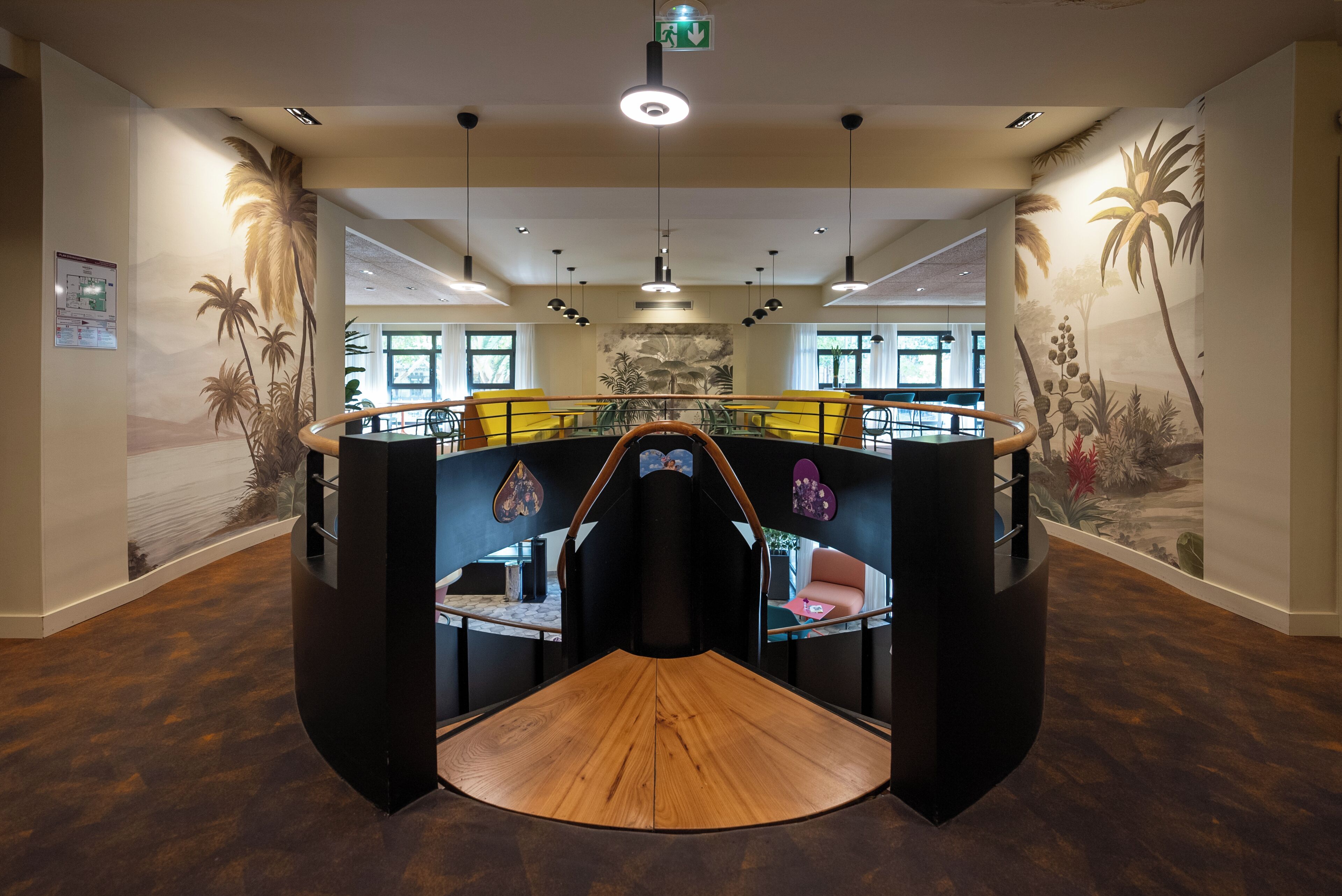Un intérieur moderne avec une plateforme centrale en bois, des rampes noires et une fresque représentant des plantes tropicales.