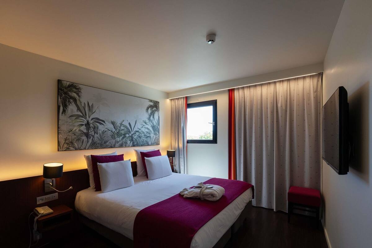 A hotel room with a bed, a painting, a window with curtains, a bedside lamp, and a television.