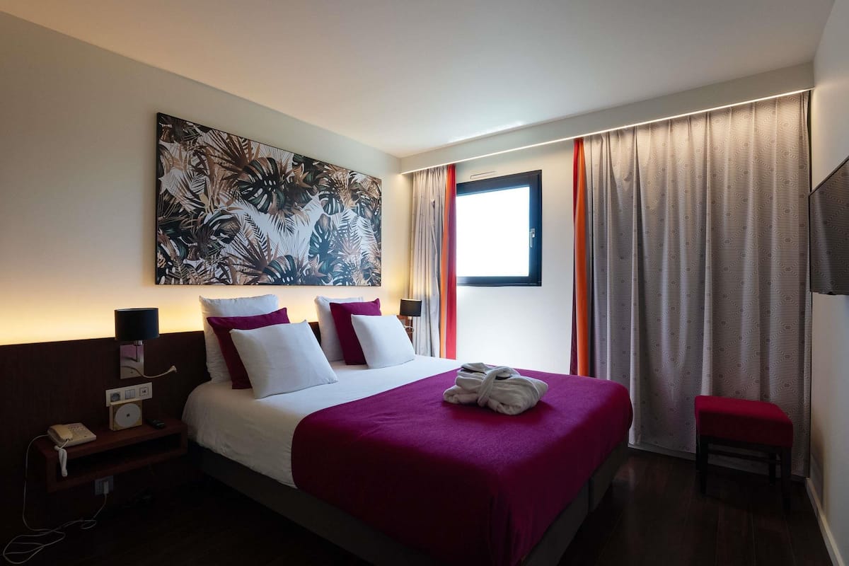 A hotel room with a large bed, a nightstand with a phone, a painting on the wall, a window with curtains, and a TV on the wall.