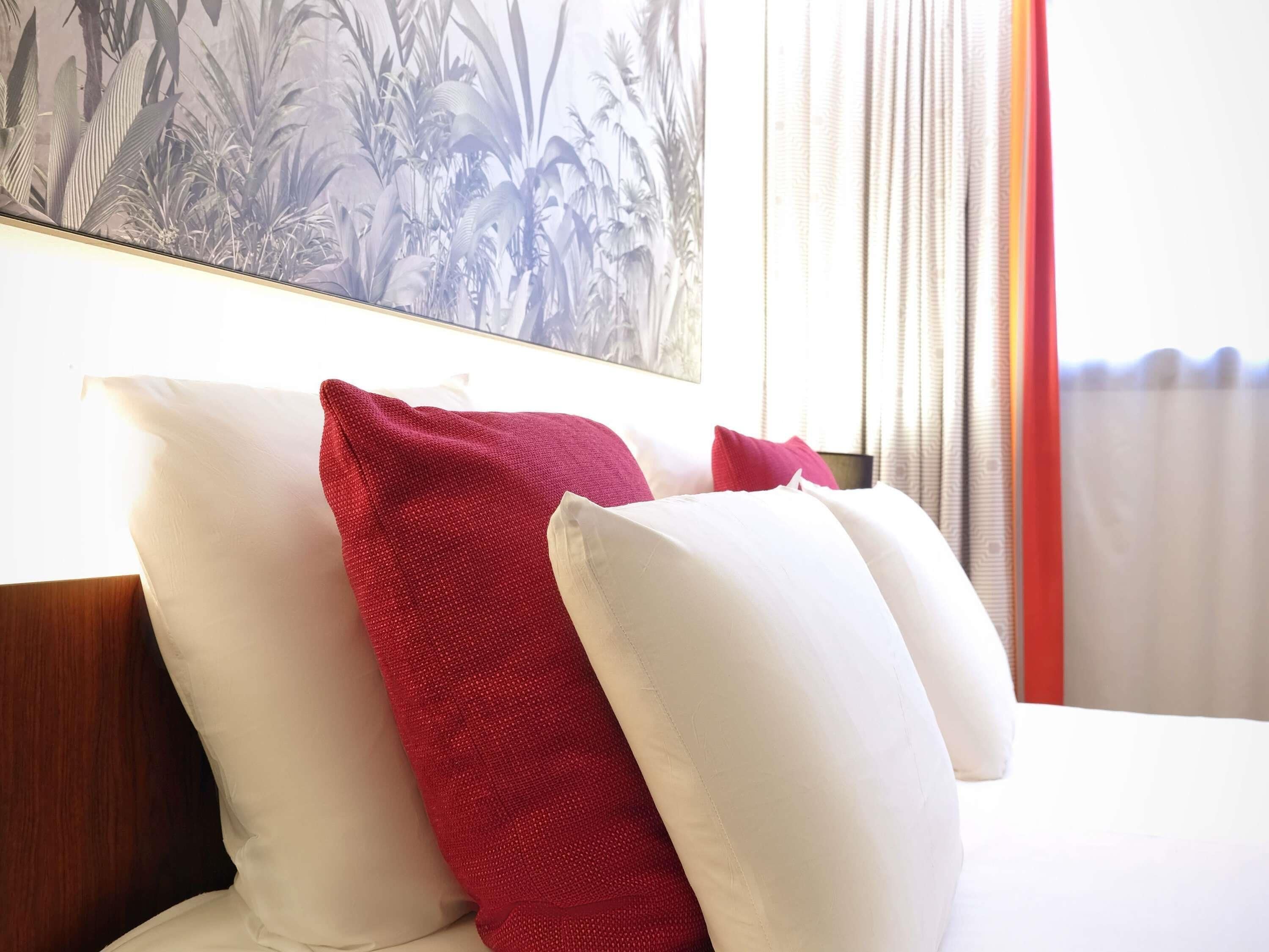 A bed with white and red pillows, a wooden headboard, and a wall with a plant mural.