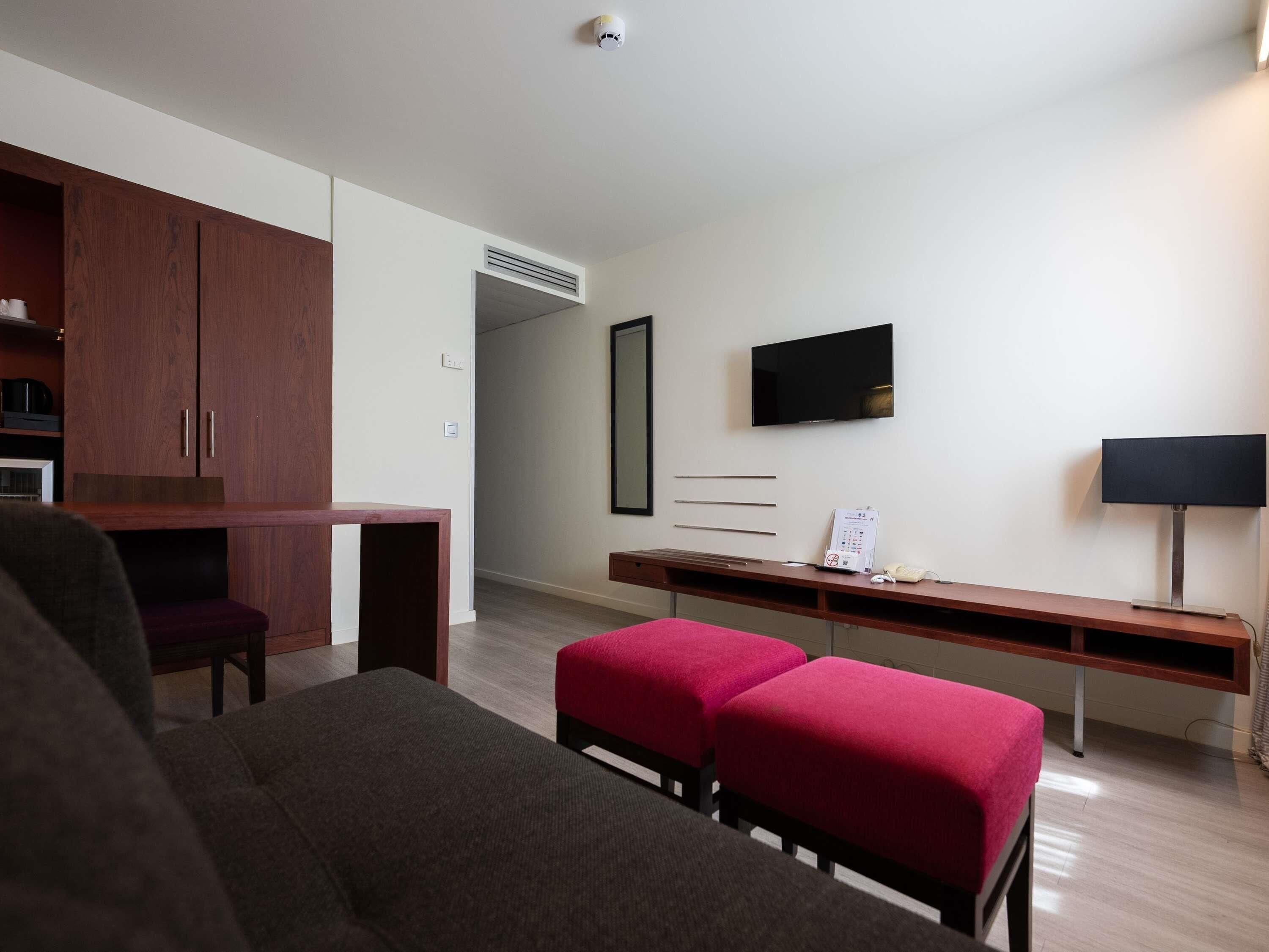 Una habitación de hotel moderna con un escritorio de madera, televisor, sofá y un reposapiés rojo.