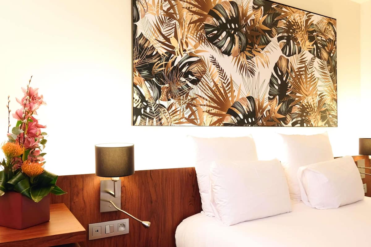 A hotel room with a bed, white pillows, a nightstand with a lamp, a wall-mounted power outlet, and a large tropical leaf-patterned painting.
