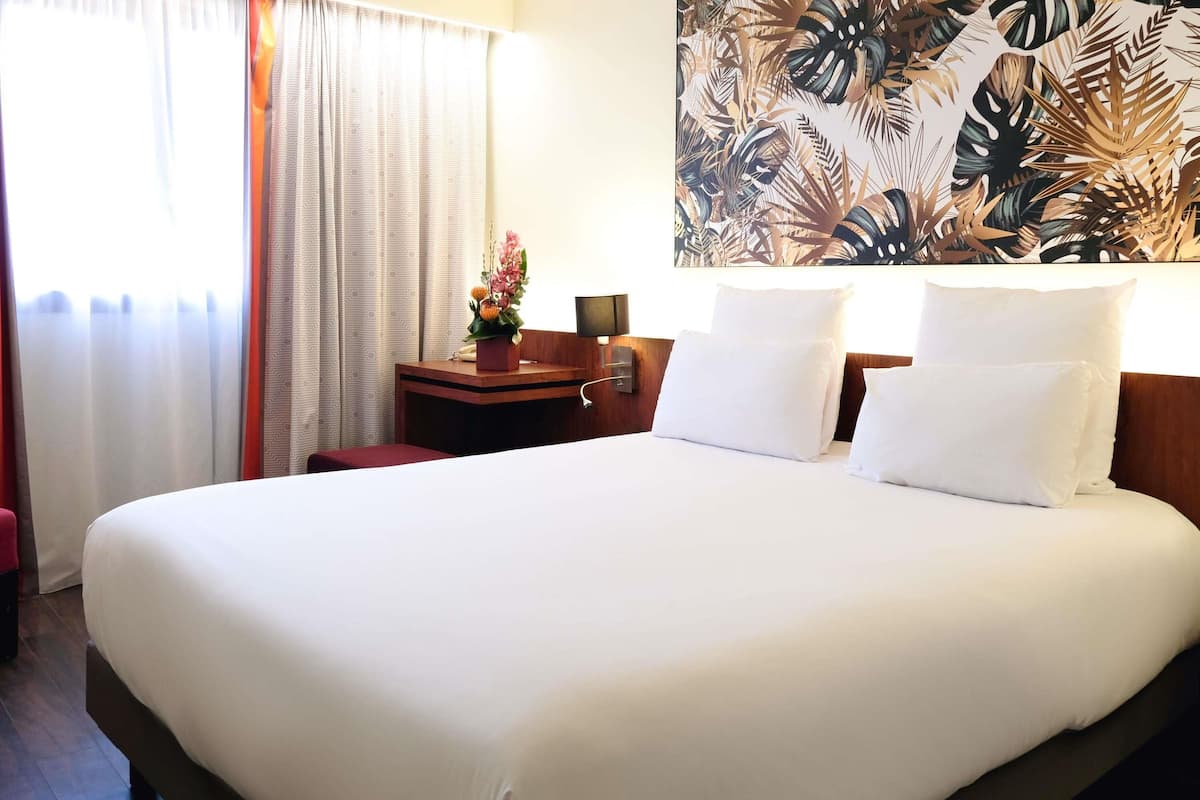 A hotel room with a large bed, a wooden nightstand, a small table with a vase of flowers, and a tropical leaf-patterned wall hanging.