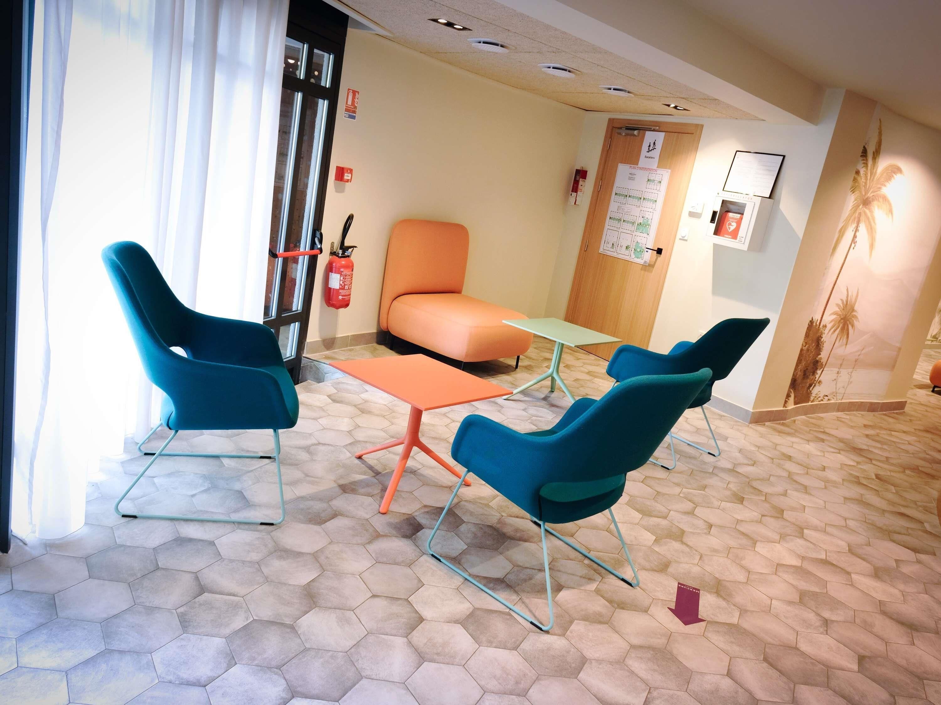 A hotel room with a hexagonal tiled floor, a teal armchair, an orange sofa, a small table, and a fire extinguisher on the wall.