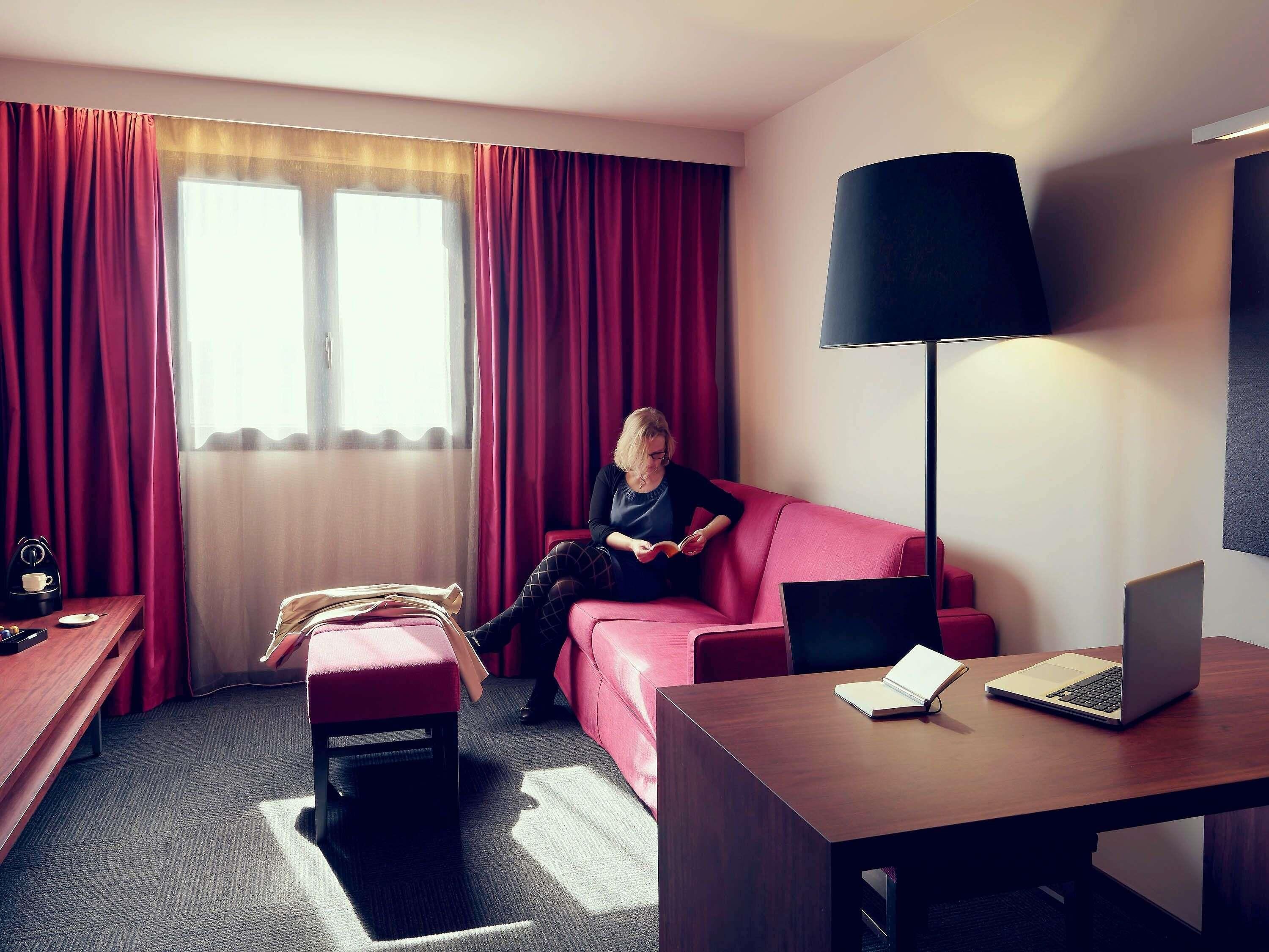 Una habitación de hotel con un sofá rojo, un escritorio de madera con una computadora portátil, una lámpara de pie y una ventana con cortinas.