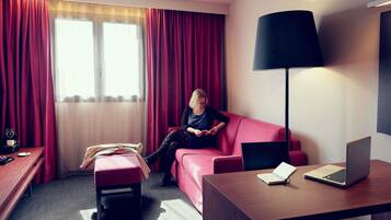 Una habitación de hotel con un sofá rojo, un escritorio de madera con una computadora portátil, una lámpara de pie y una ventana con cortinas.