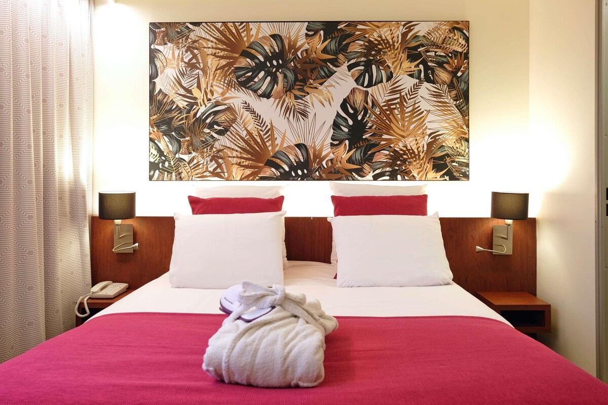 A hotel room with a bed, two bedside tables, a telephone, a towel, and a large tropical leaf-patterned artwork on the wall.