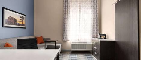A modern hotel room with a sofa, a dining table, a desk, a large window with curtains, and a wardrobe.
