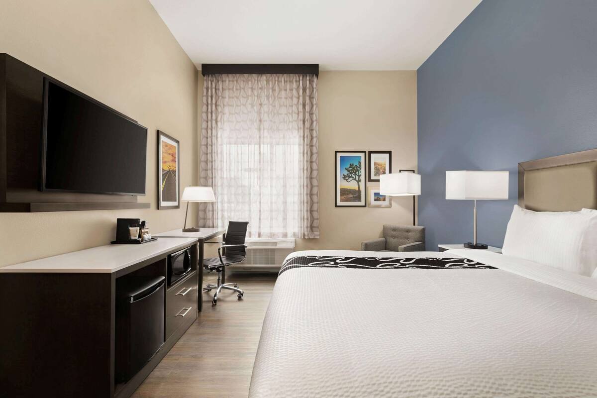 A hotel room with a bed, a desk with a chair, a TV, and a window with curtains.
