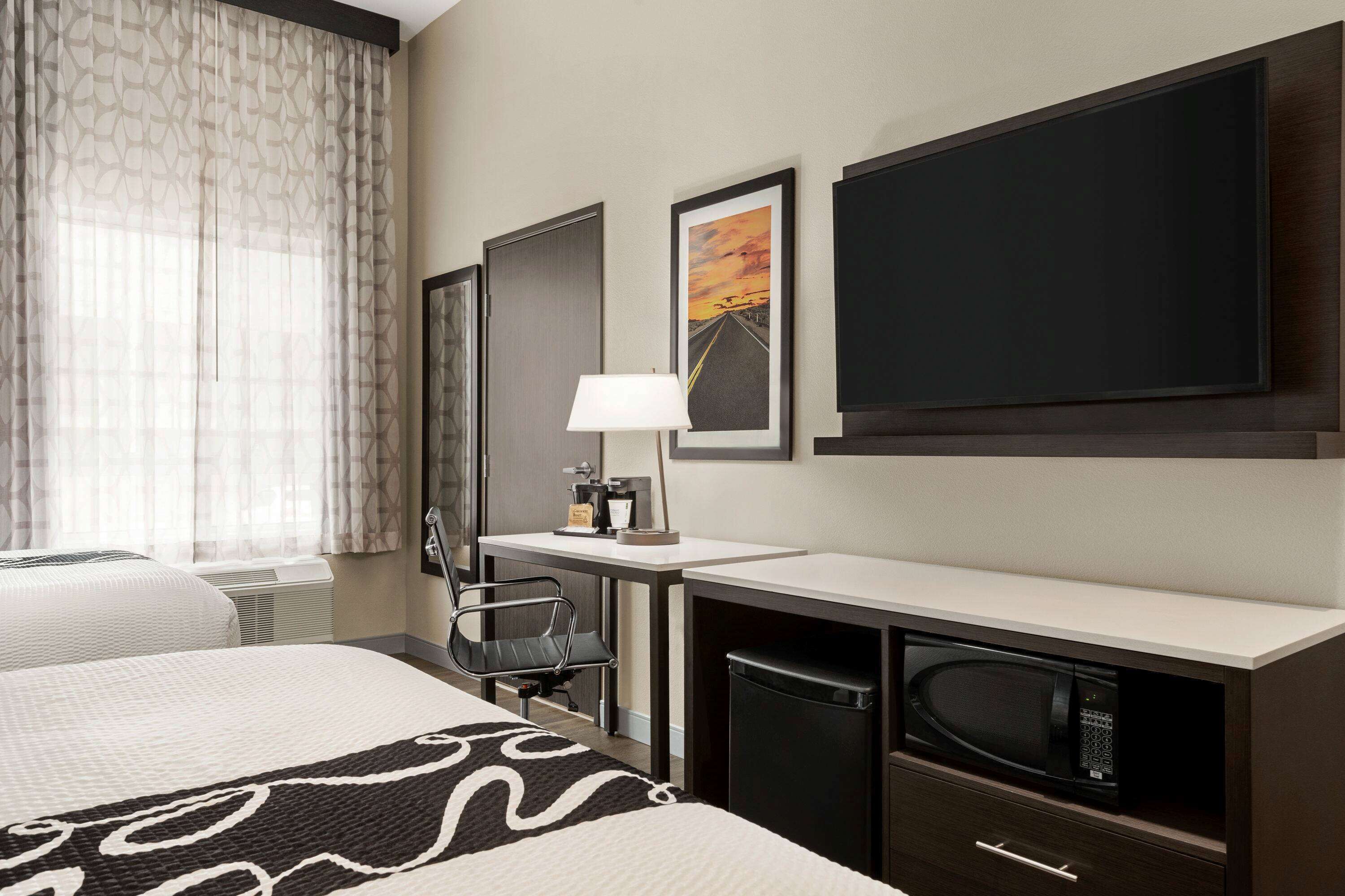 A hotel room with a bed, a desk with a lamp, a chair, a microwave, and a large framed picture on the wall.