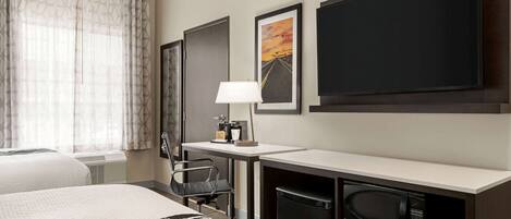 A hotel room with a bed, a desk with a lamp, a chair, a microwave, and a large framed picture on the wall.