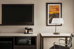 Habitación de hotel con una televisión de pantalla plana grande, un cuadro enmarcado de una puesta de sol, una estación de café, un microondas y una silla moderna.
