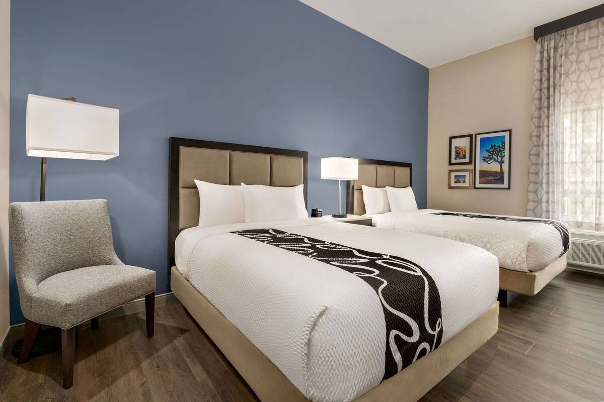 A hotel room with two beds, a chair, a lamp, and two framed pictures on the wall.