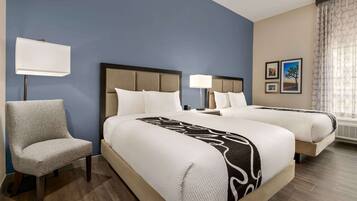 A hotel room with two beds, a chair, a lamp, and two framed pictures on the wall.