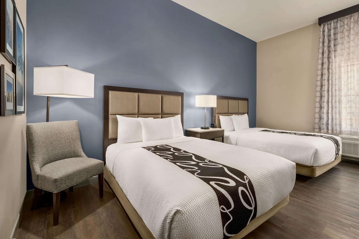 A hotel room with two beds, a chair, a nightstand with a lamp, and a window with curtains.