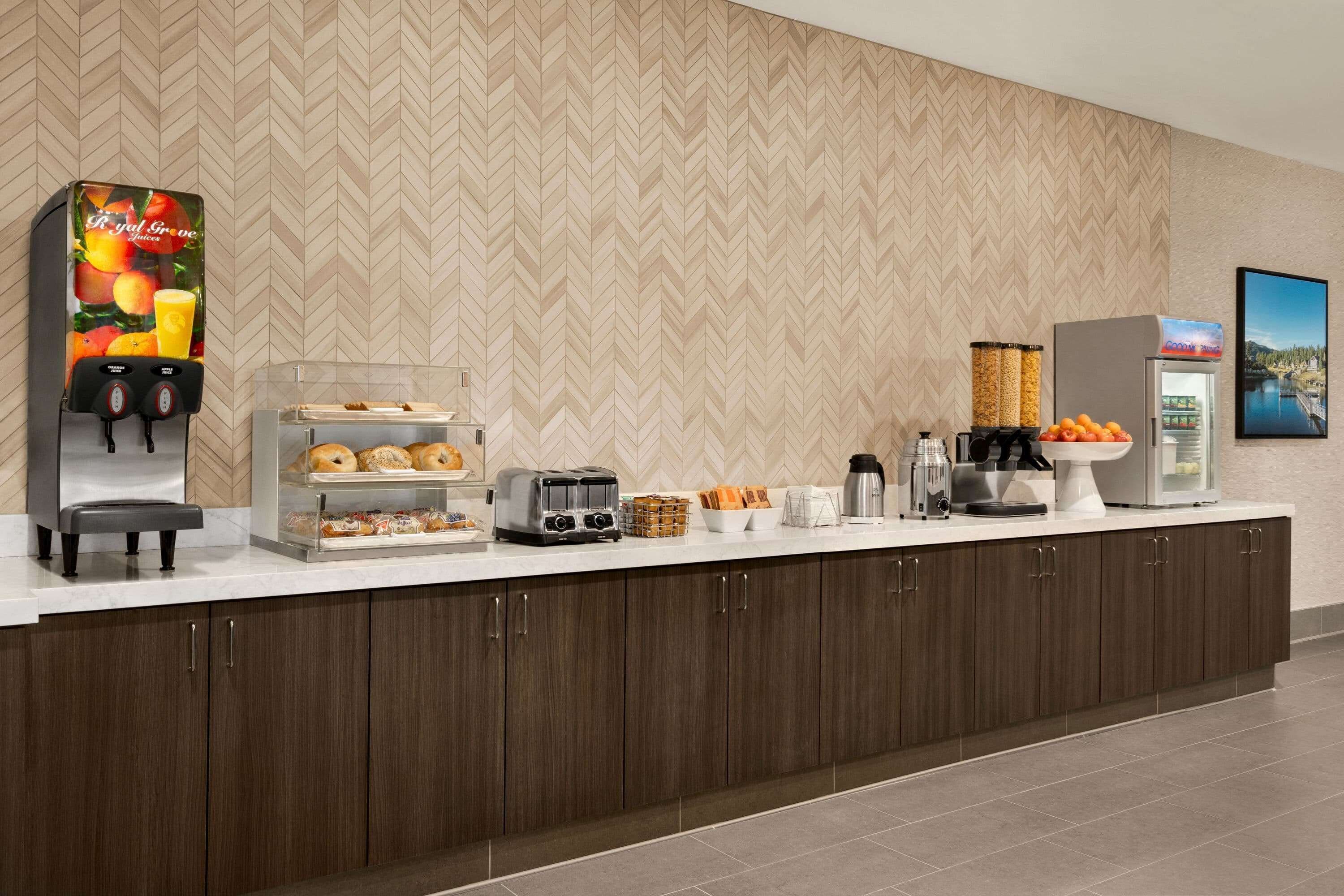 A hotel breakfast area with a coffee machine, toaster, fruit display, and a refrigerator.