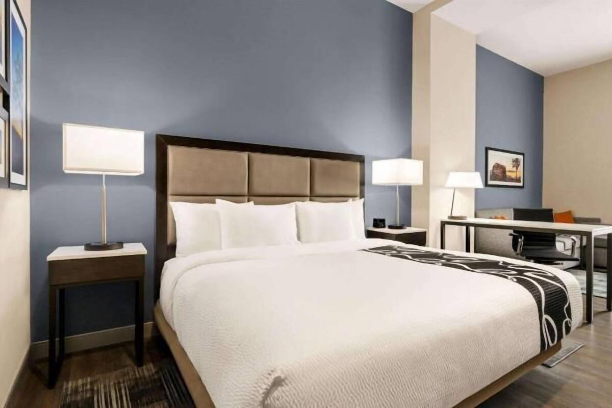 A hotel room with a large bed, two bedside tables with lamps, a desk with a chair, and a window with curtains.